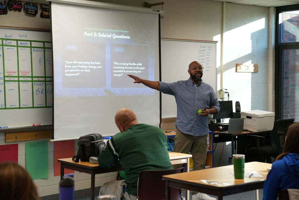 A presenter with glasses and a beard stands in front of a projection screen gesturing toward slide titled "Part 2: Debrief Questions," which features discussion prompts about resiliency and adaptability during a professional development session.