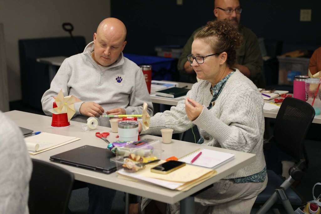 Two educators sit side-by-side at a desk assembling hands-on project materials, including paper pinwheels, string, and cups, with laptops and supplies spread across the table.