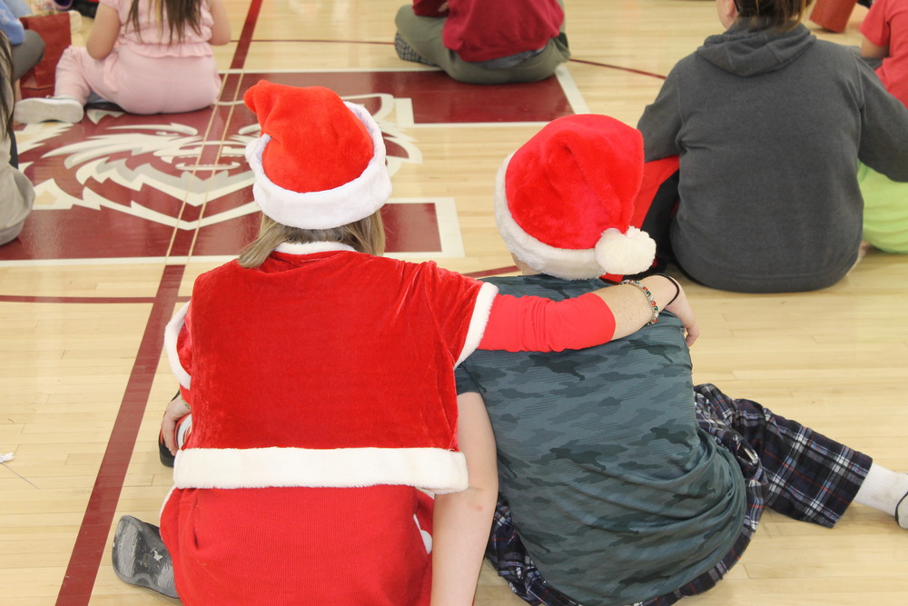 students sitting on gym floor in holiday clothes, one with arm around the other