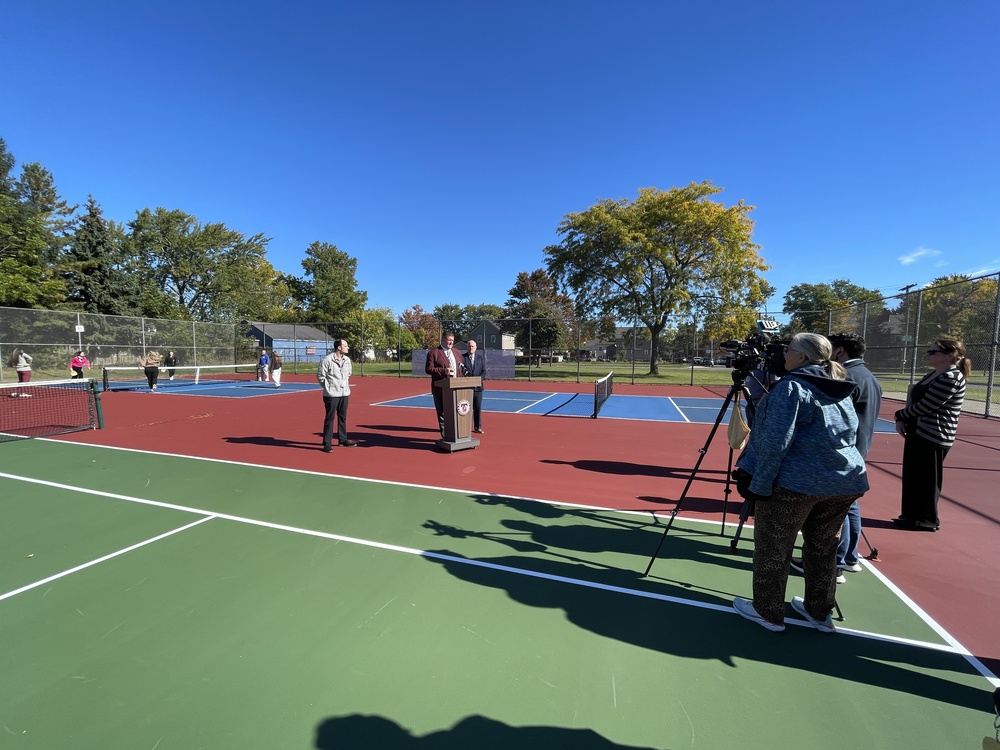 wide shot of press conference with cameras in foreground, podium and speakers in center and pickleball players in distance