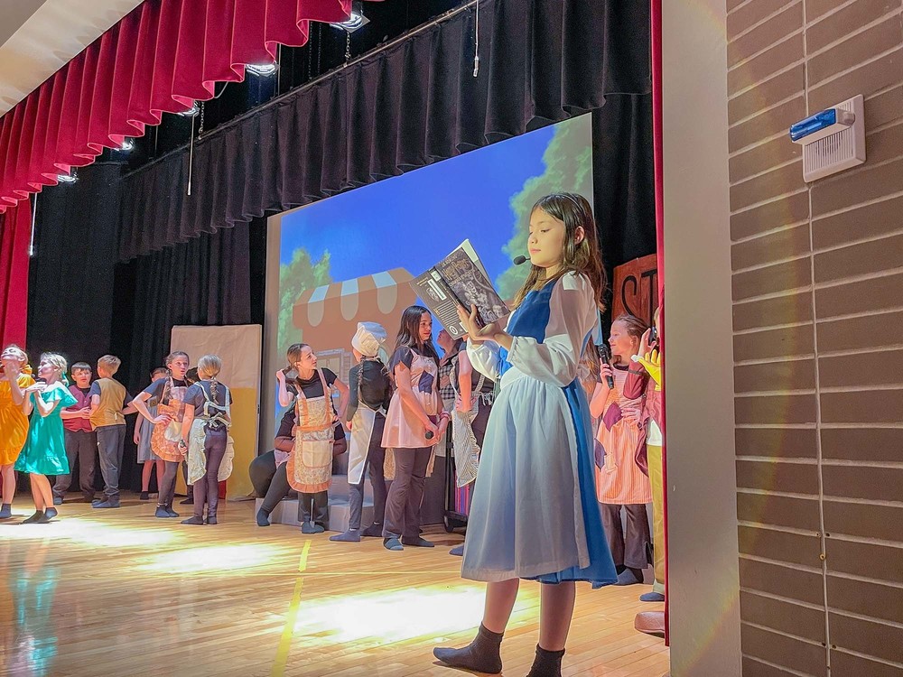 belle reads in the foreground while the rest of cast interacts behind her on the stage
