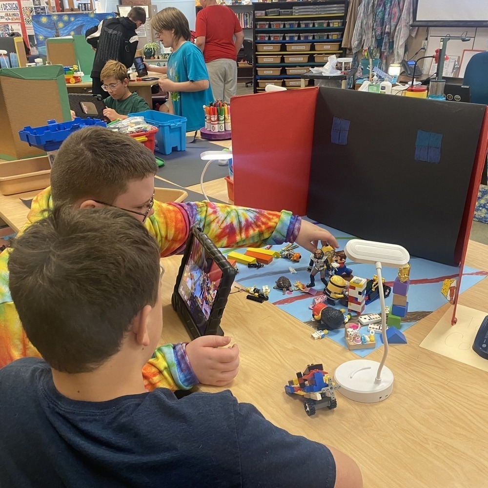 boy wearing tie dye shirt and boy wearing black t shirt sit at a table using Lego figurines to create a scene for their stop-motion movie