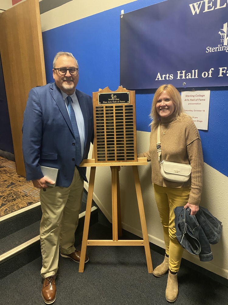 Mr Harrell wearing glasses, a light blue shirt, blue tie and sport coat, and khakis; Mrs. Harrell wearing a brown sweater and yellow pants; a wooden easel with metal name plaques stands between them