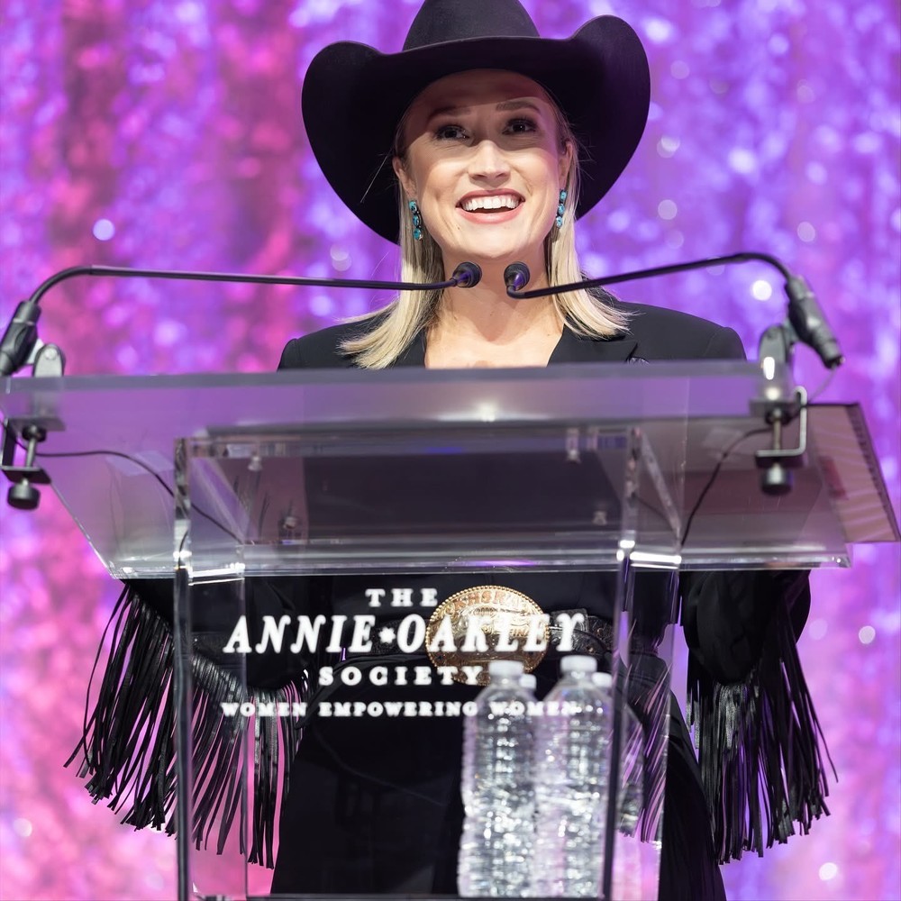 blonde woman wearing black leather jacket and cowboy hat, standing at a glass lectern