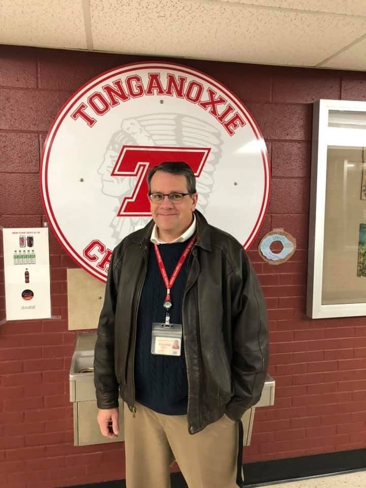 Mr. Gifford wearing navy sweater, black leather jacket, and khakis, standing in front of a red brick wall and circular Tonganoxie Chieftains sign