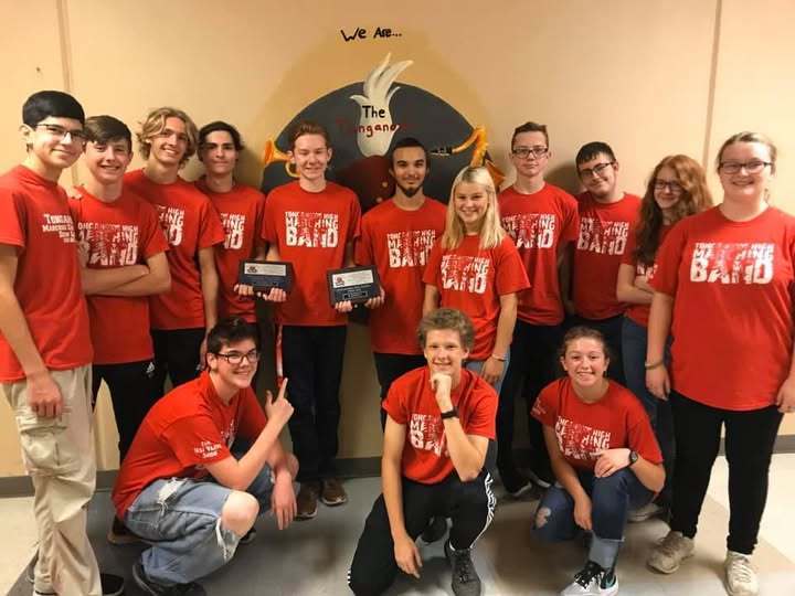 14 band and drumline members wearing matching red shirts, two in the middle hold award plaques for their performances