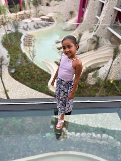 Ana Franco stands on a glass balcony of a hotel wearing a purple tank top and floral skirt