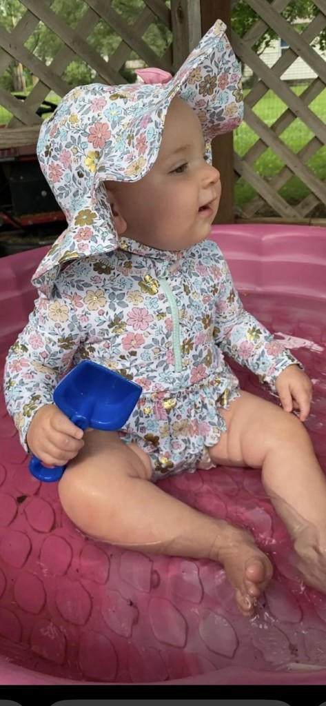 Baby Willow sitting in a pink kids pool, wearing a flowered swimsuit and hat, holding a blue shovel