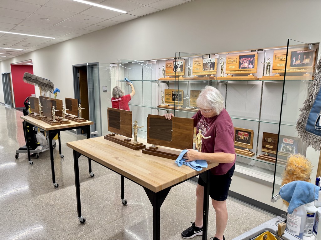two female custodians dusting trophies in a wall trophy case