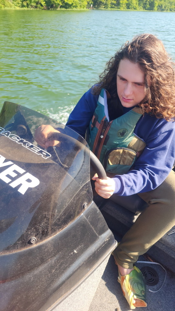 Sawyer Vestal wearing long sleeved blue shirt and green life jacket, sitting in the driver's seat of a fishing boat