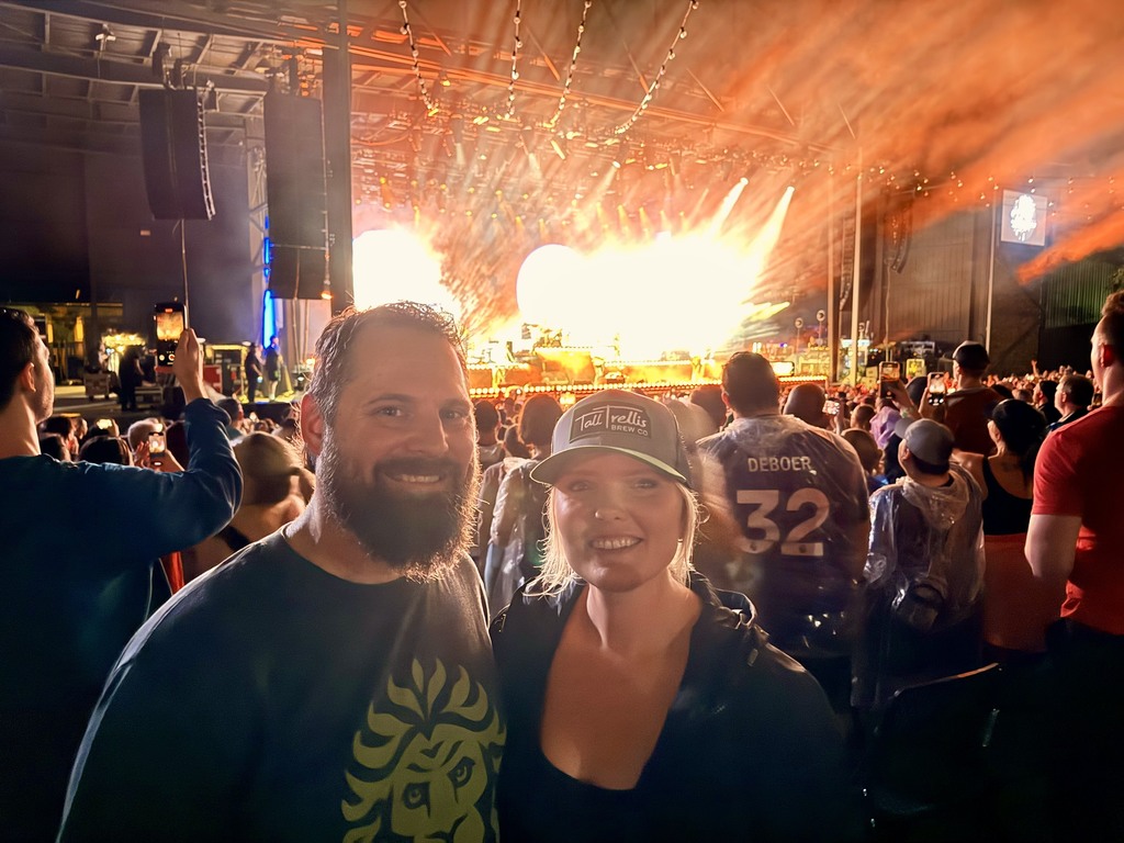 Kelly Griffiths wearing a baseball cap and dark colored top, standing next to her husband wearing a dark colored t shirt, with the Azura Amphitheater stage in the background