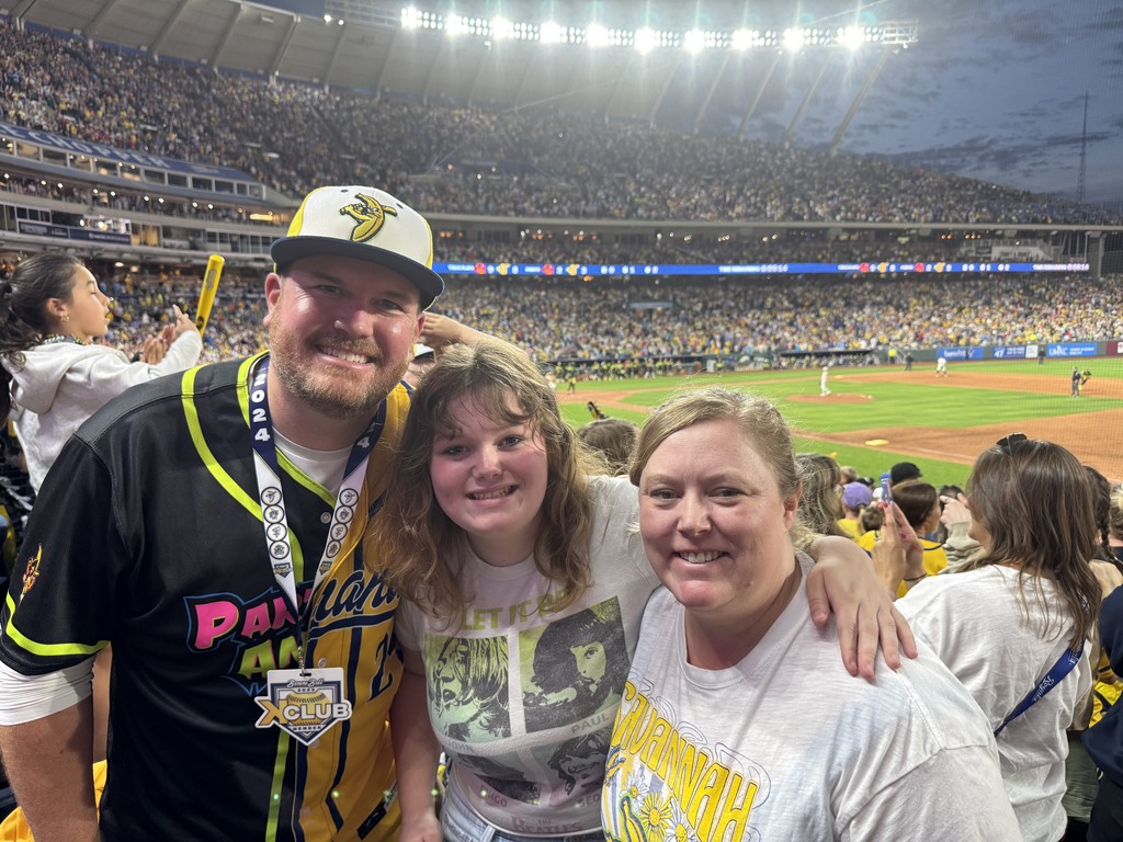 Matt Bowers, wife and daughter standing together at a baseball game, the lights of the stadium shine brightly along the top of the image