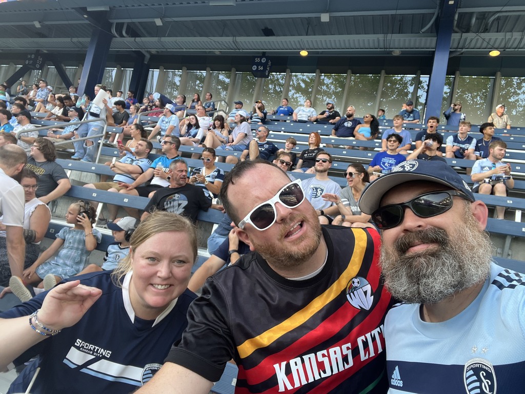 Matt Bowers wearing white sunglasses, flanked on either side by his wife and a friend, taking a selfie together in the blue seats of a soccer stadium