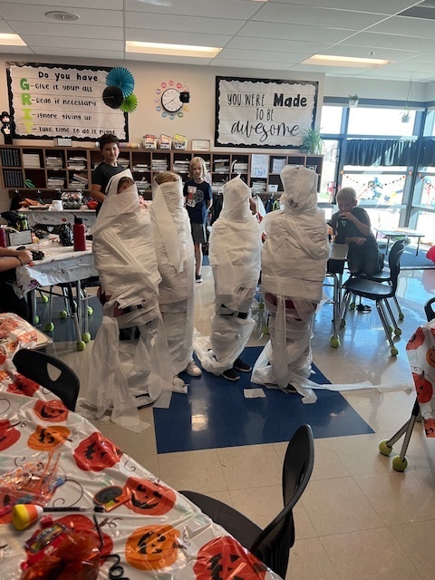 four fourth grade students wrapped in toilet paper stand together in a fourth grade classroom