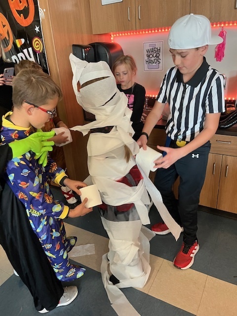 fourth grade students work together to wind toilet paper rolls around a student to create a mummy