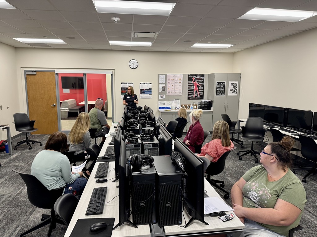 seven adults seated at computer stations, tonya phillips wearing pants and black polo stands at front of room speaking