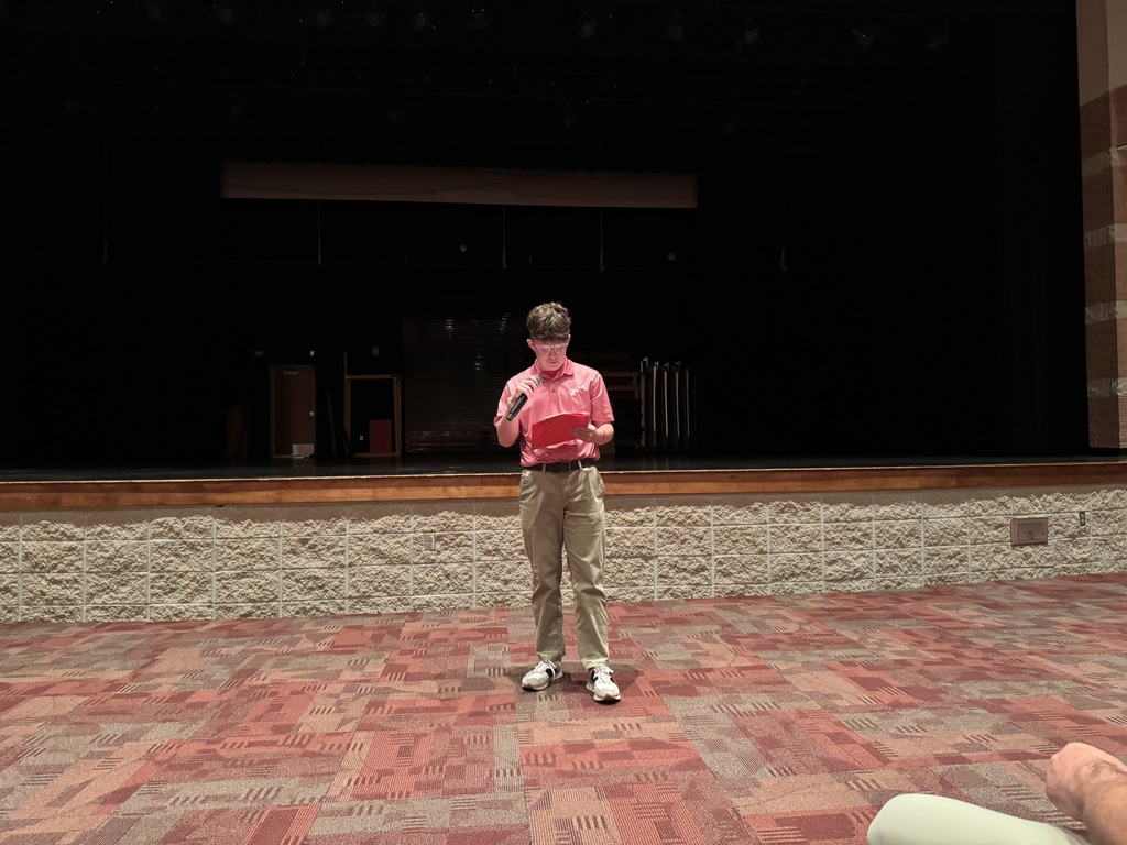 Wesley stands at the front of the auditorium holding a mic and a piece of paper