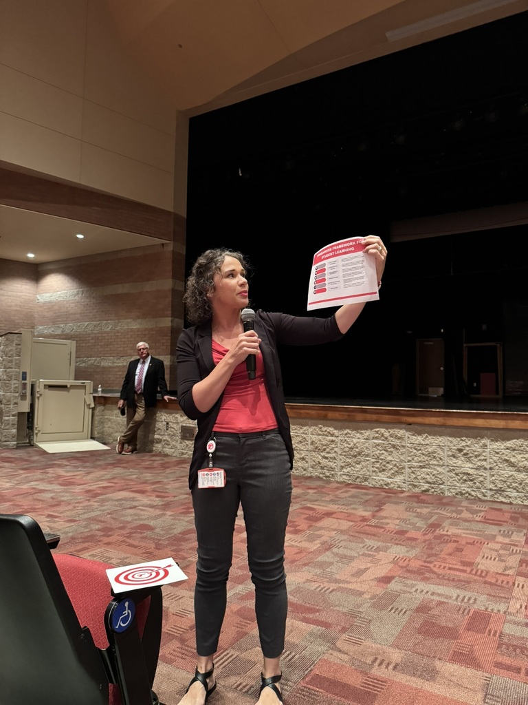 Brandy Taylor wearing red shirt black pants black blazer, holding a microphone in her right hand and a piece of paper in her left