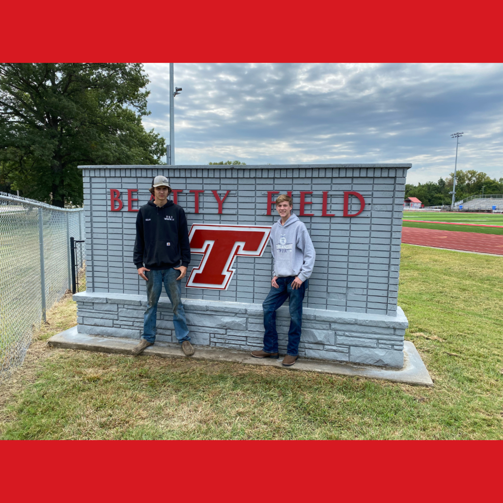 two high school boys wearing hoodies and jeans, standing in front of a gray brick Beatty Field sign with a red and white welded T on it