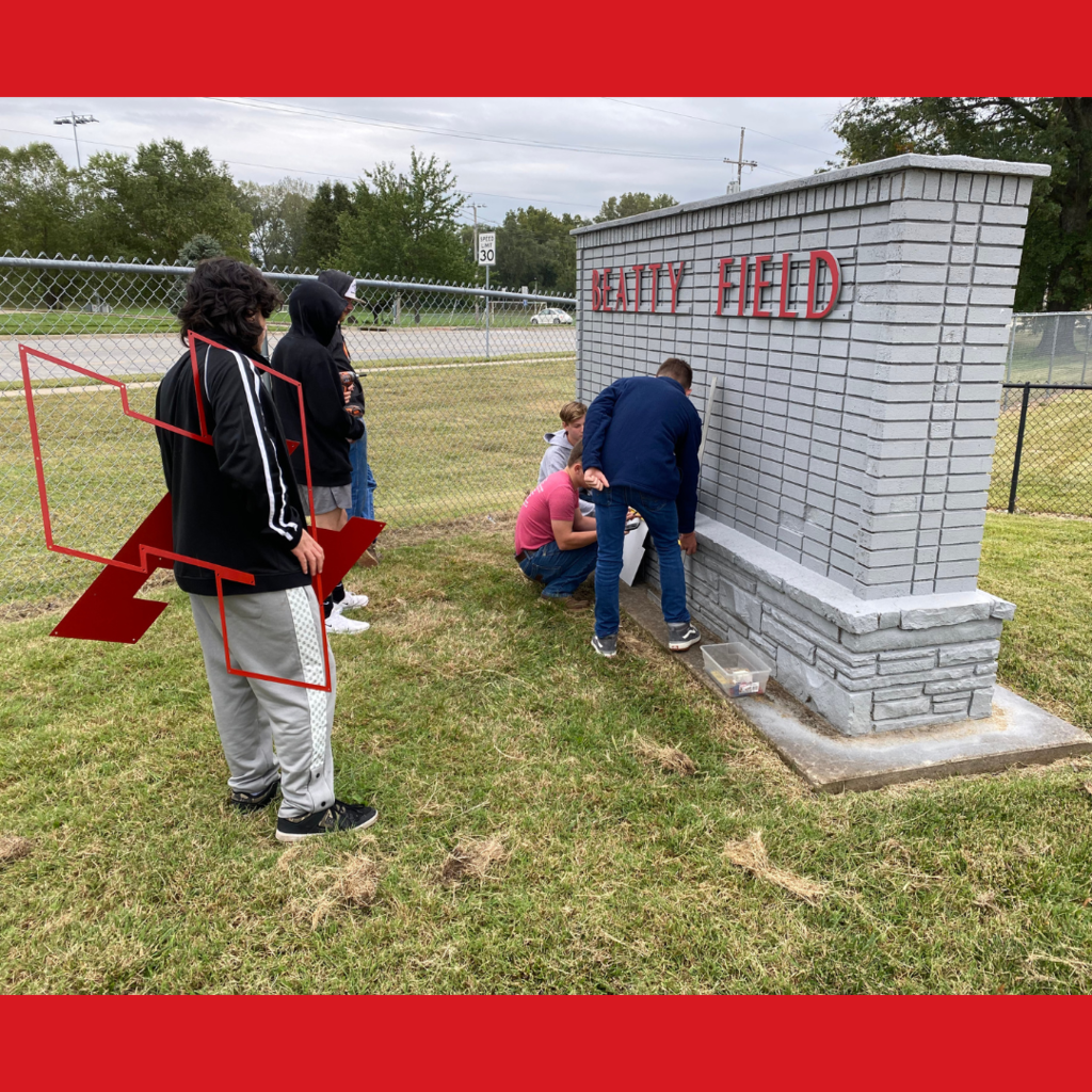 three males working on a gray brick sign while three others look on