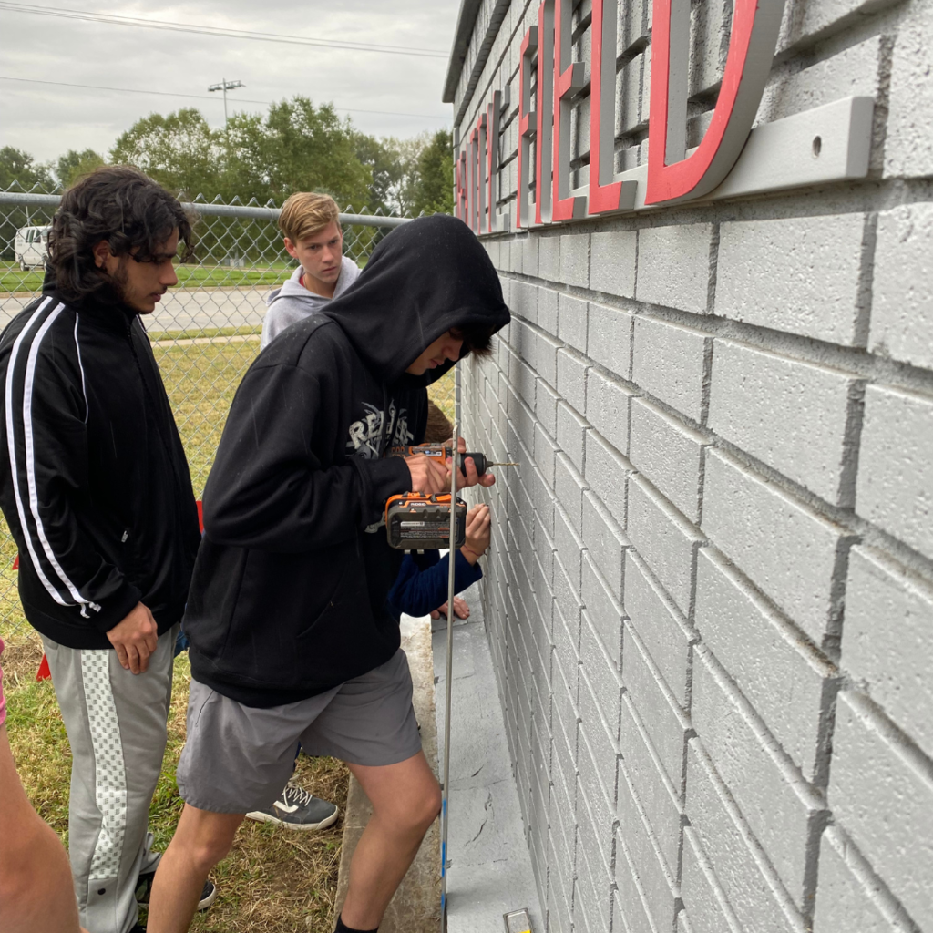male wearing shorts and black hoodie screws brackets into gray brick wall