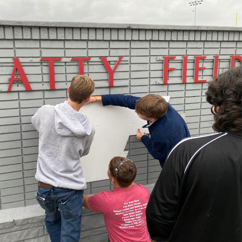 three males hold up the white Tongie T against the gray brick sign