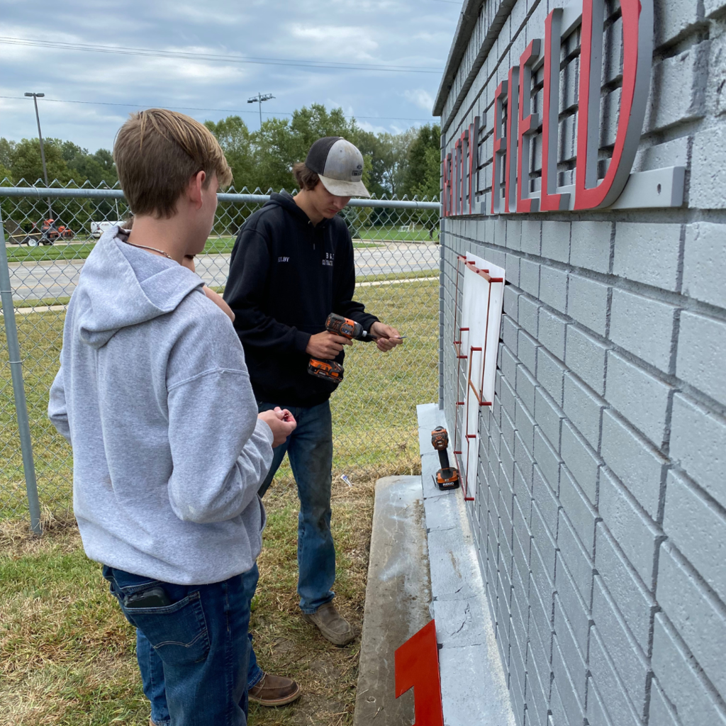 three males, one holding a drill, work on attaching a metal T to the gray brick wall