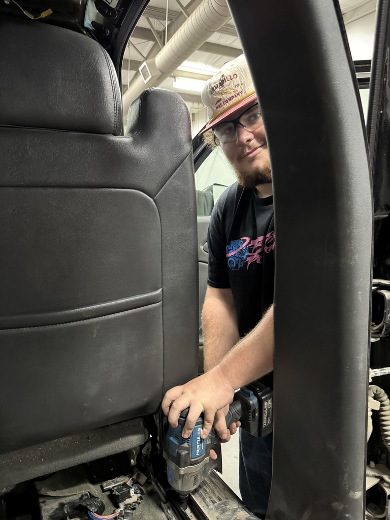 student in black t shirt and white hat uses a power drill to remove a bolt from a truck seat