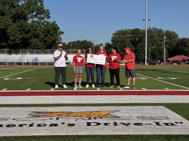 Bank employees and superintendent wearing red shirts standing on the football field, accepting a check