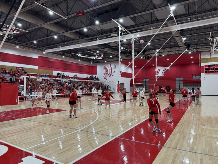 High school girls wearing red or white jerseys playing volleyball in a high school gymnasium