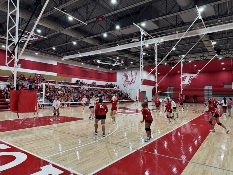 High school girls wearing red or white jerseys playing volleyball in a high school gymnasium