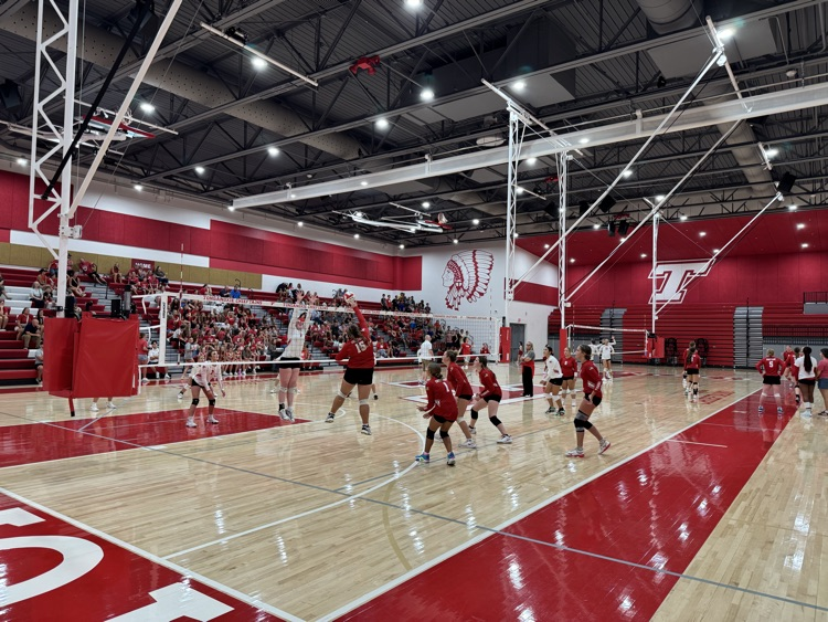 High school girls wearing red or white jerseys playing volleyball in a high school gymnasium