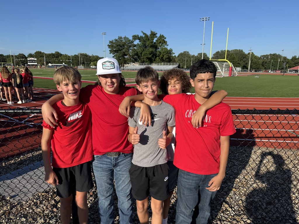 five boys stand with their arms around each others' shoulders, learning up against a black chain link fence