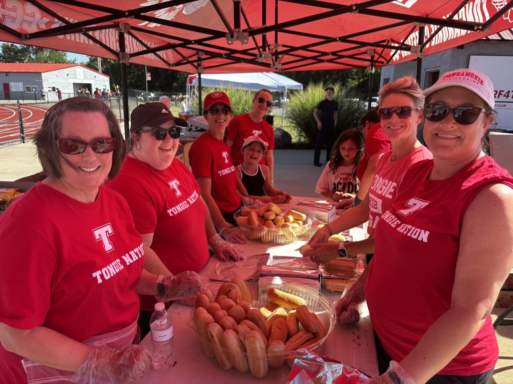 district administrators wearing red t shirts and plastic gloves stand before a table of hot dogs buns, ready to serve hot dogs