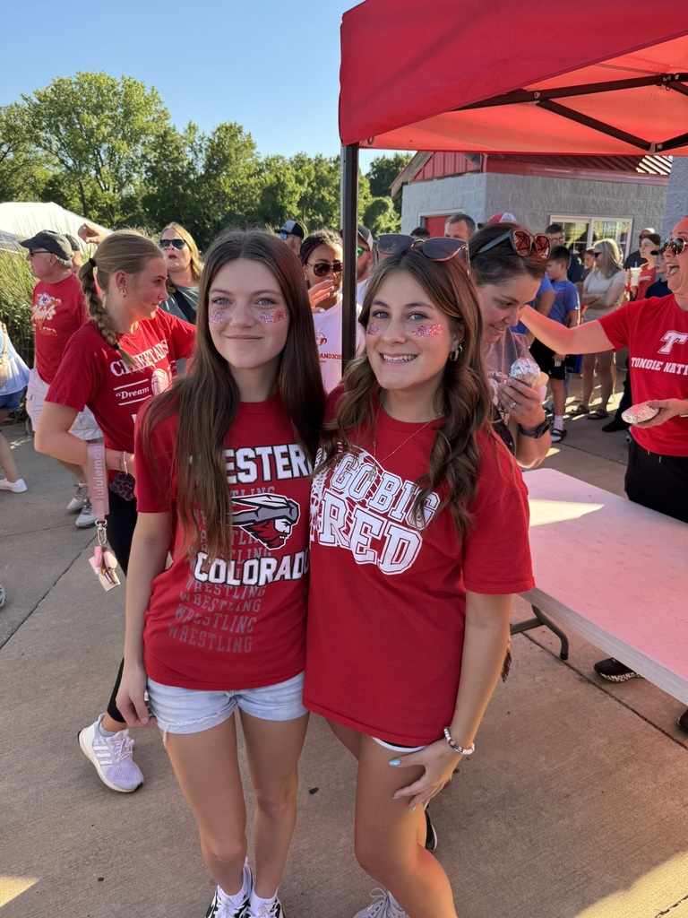 two high school girls wearing red t shirts and shorts, standing together in front of a white table and red tent