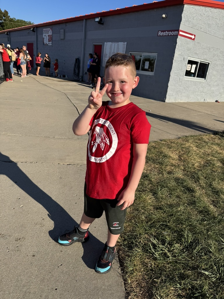 an elementary school boy wearing red t shirt and black pants, giving the two fingers peace sign