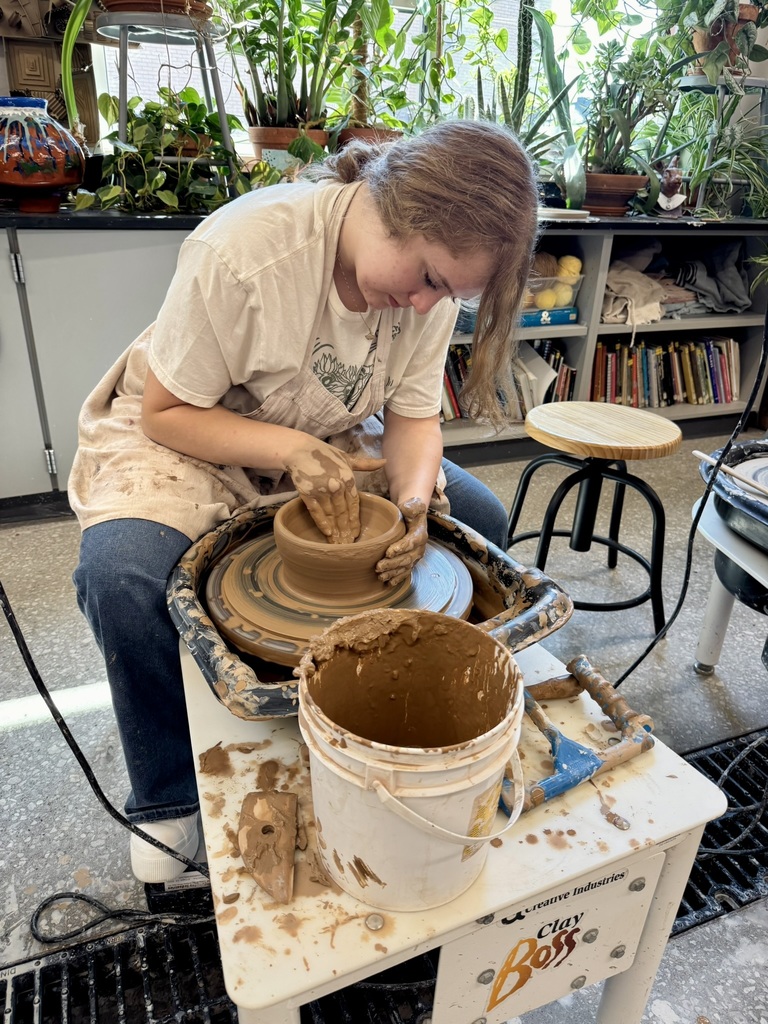 Ava Bond wearing jeans, a white shirt, and an apron, making a clay pot on a wheel