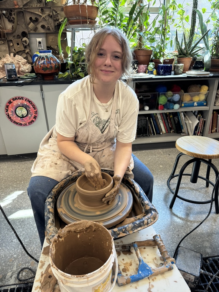 Ava Bond wearing jeans, a white shirt, and an apron, making a clay pot on a wheel