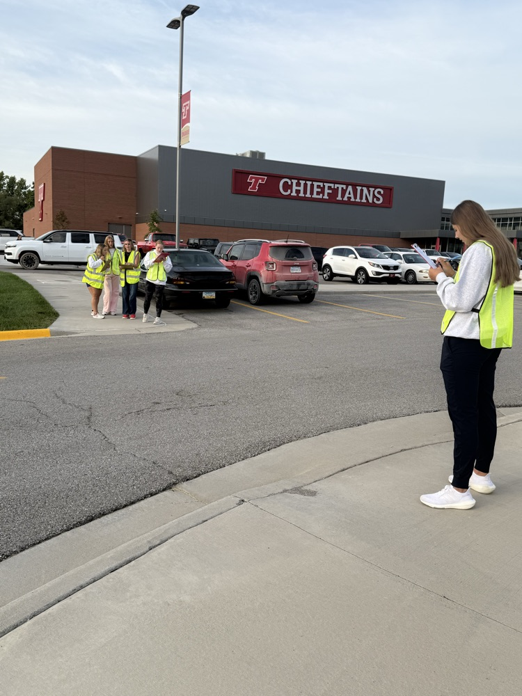 students standing in the parking lot of a high school wearing high vis vests and holding clipboards