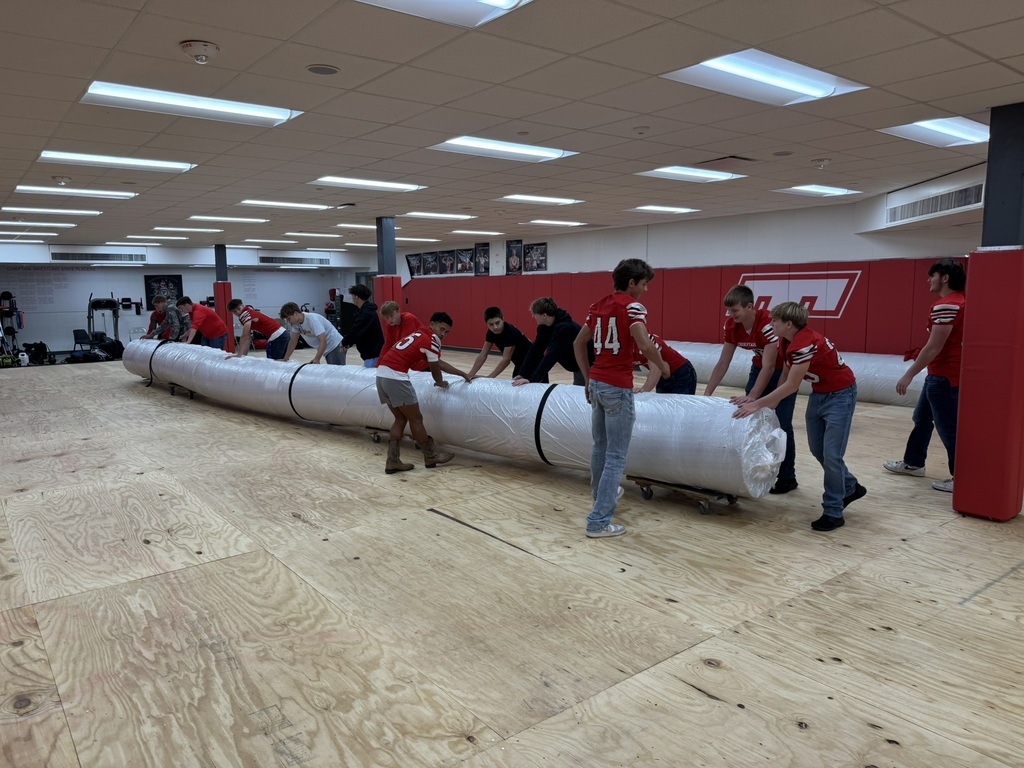 high school boys wearing red football jerseys move wrestling mats into the district administrative center