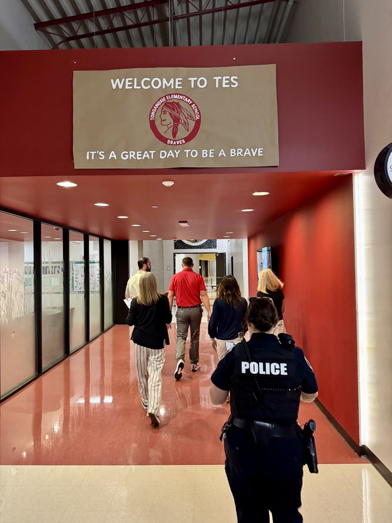 TES safety team members walk down the hallway at an elementary school: six adults, one in a police officer uniform that says POLICE on the back