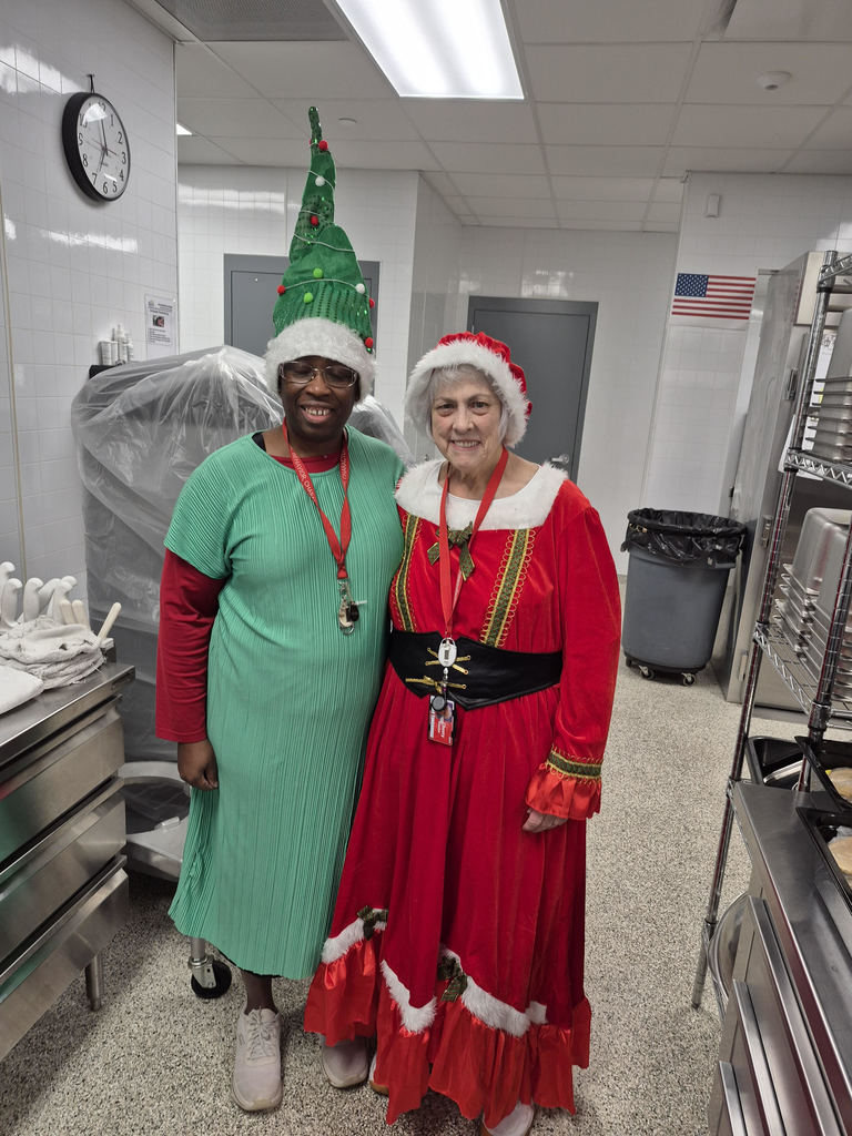 two women standing in a high school cafeteria kitchen; one on the left wears a green dress with red sleeves and a Christmas tree hat; the one on the right wears a red Mrs. Claus dress and Santa hat