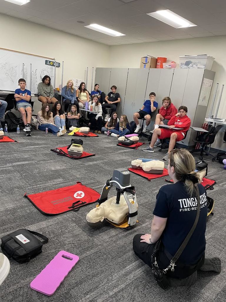 students look on at a female firefighter demonstrates the use of Lucas 3, a device that performs chest compressions on a mannequin