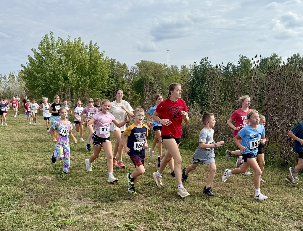 elementary runners run along the grassy course, a row of trees is behind them