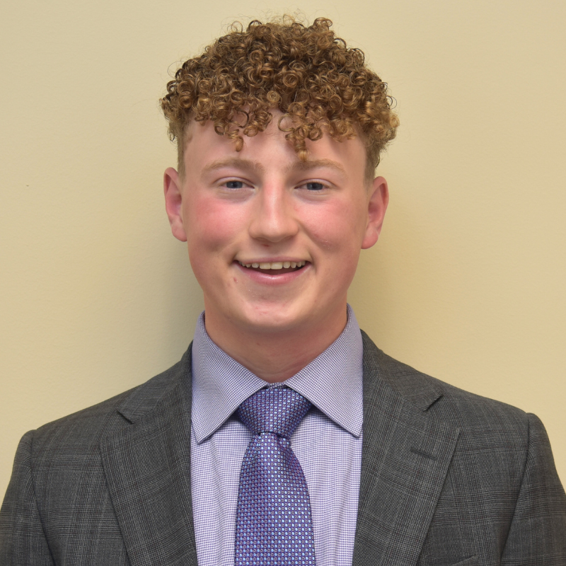 headshot of Remi Baldock wearing purple shirt and tie and gray suit jacket