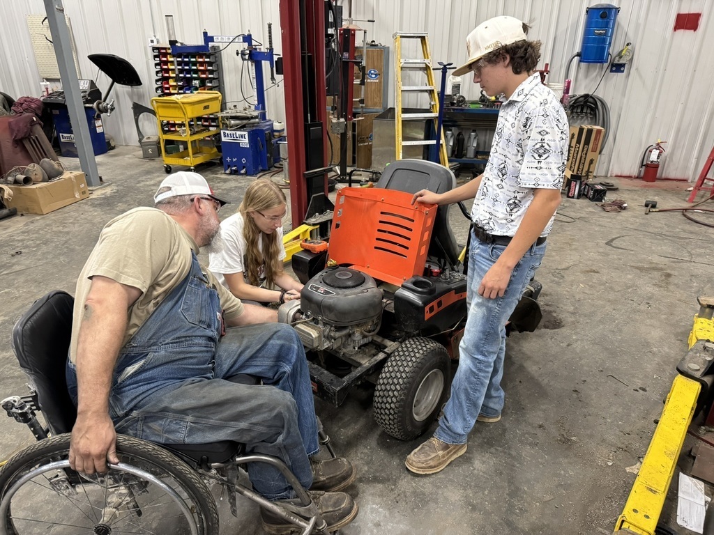 two high school students working on a riding lawn mower as Mr. Maurer looks on