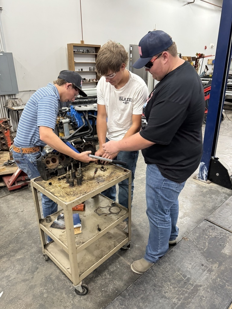 three high school boys wearing jeans and t shirts working on a motor head that sits on a rolling cart
