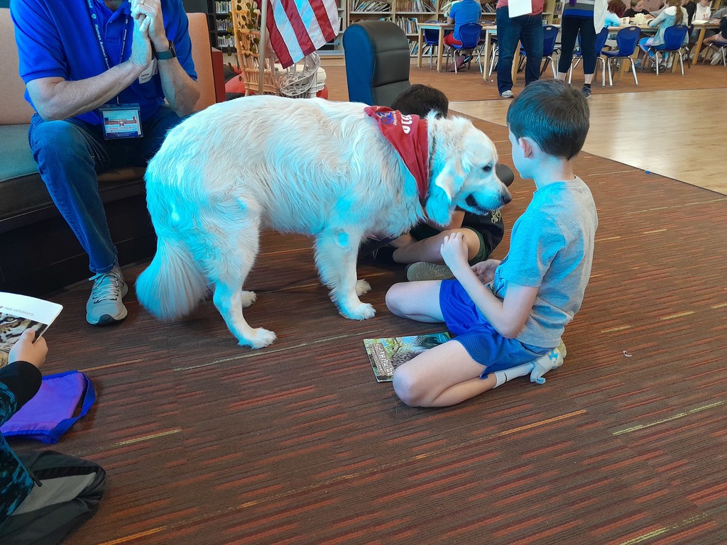 two fourth grade boys read books to a large white dog wearing a red bandana as an adult in jeans and a blue polo looks on
