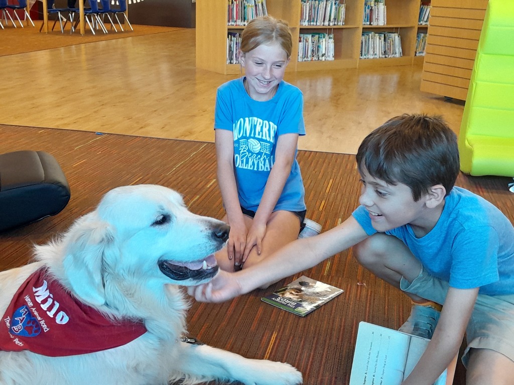 two fourth grade students read books to a large white dog wearing a red bandana