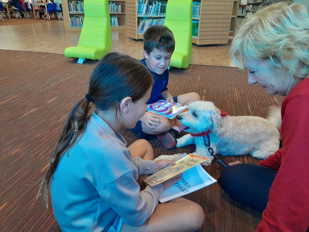 two fourth grade students read books to a white dog wearing a red collar, as a woman in a red sweater looks on
